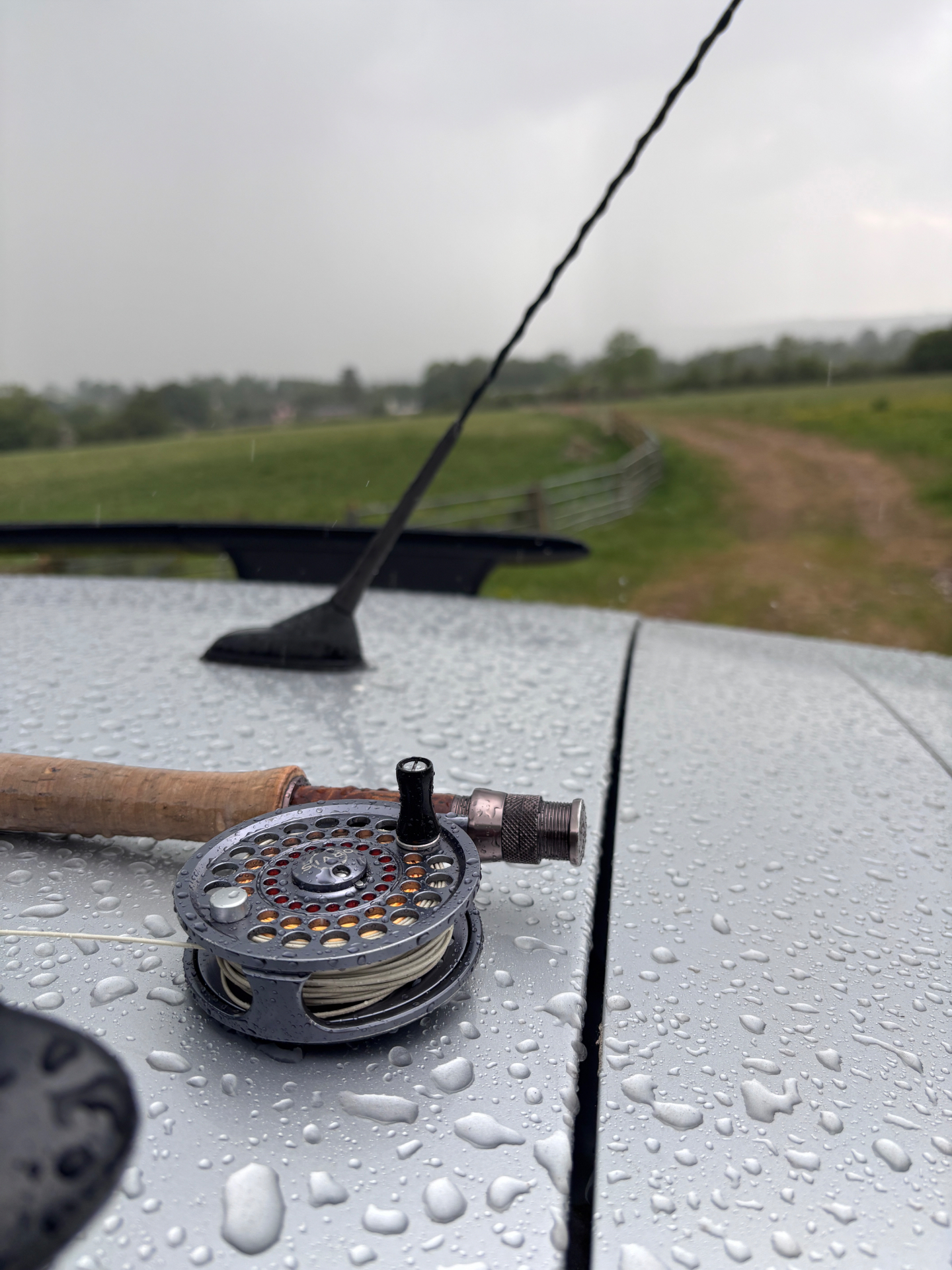 A fly rod and reel resting on a rain-spattered car bonnet, overlooking Midlands countryside